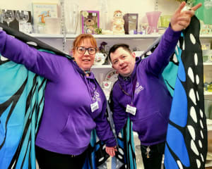 Two DEBRA team members standing in a shop wearing purple hoodies and blue butterfly wings