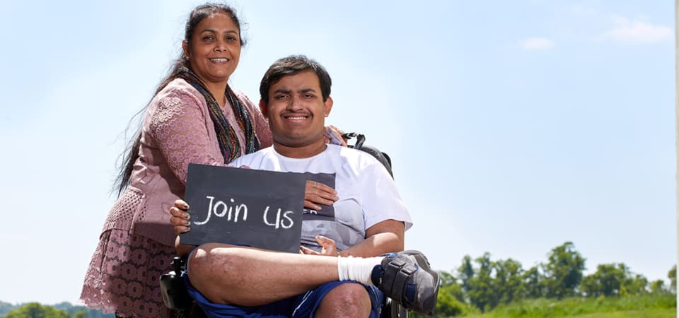 Woman hugs man in a wheelchair who is holding a chalkboard with the words Join us written