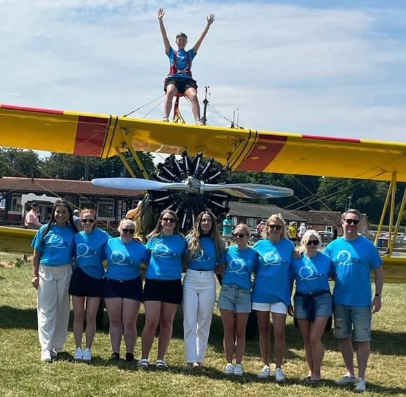 Jellybean team stand in front of a small propellor pain with one of the team strapped to the top of the plane with her arms in the air.
